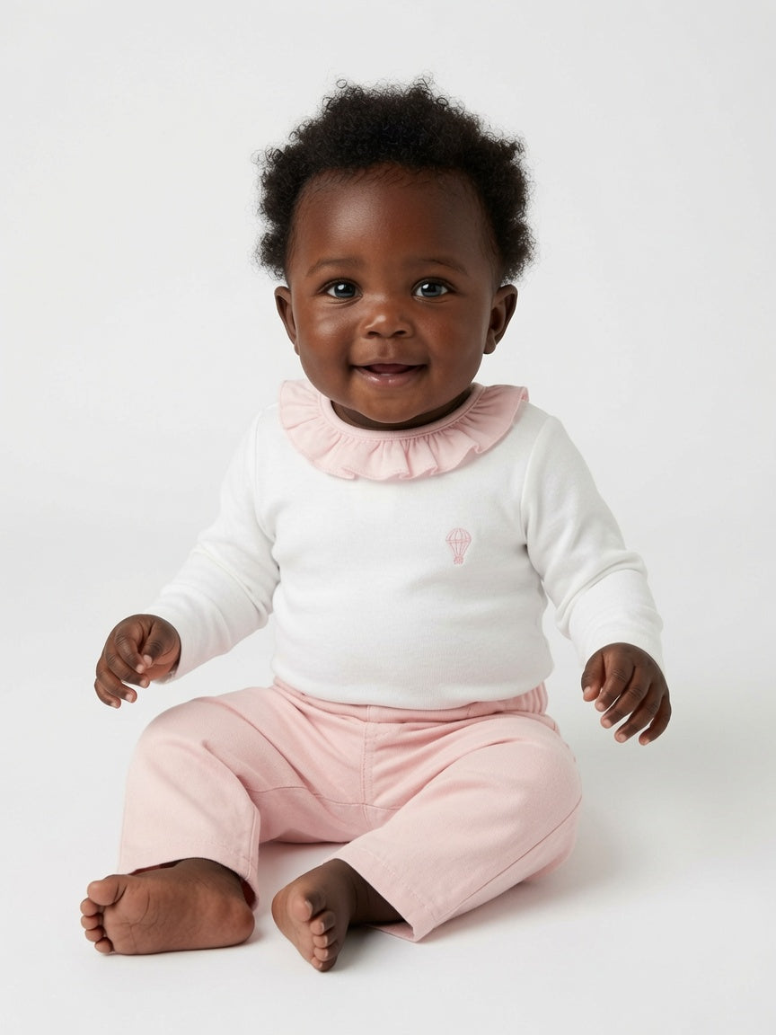 A smiling baby sitting on a white studio background, wearing a white long-sleeve top with a light pink ruffled collar and light pink pants.