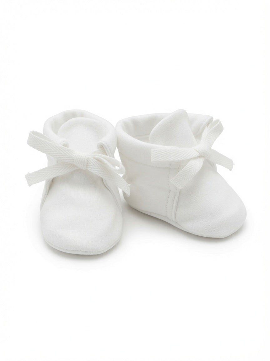 A pair of white soft fabric baby booties with matching ribbon bows, photographed on a seamless white background.