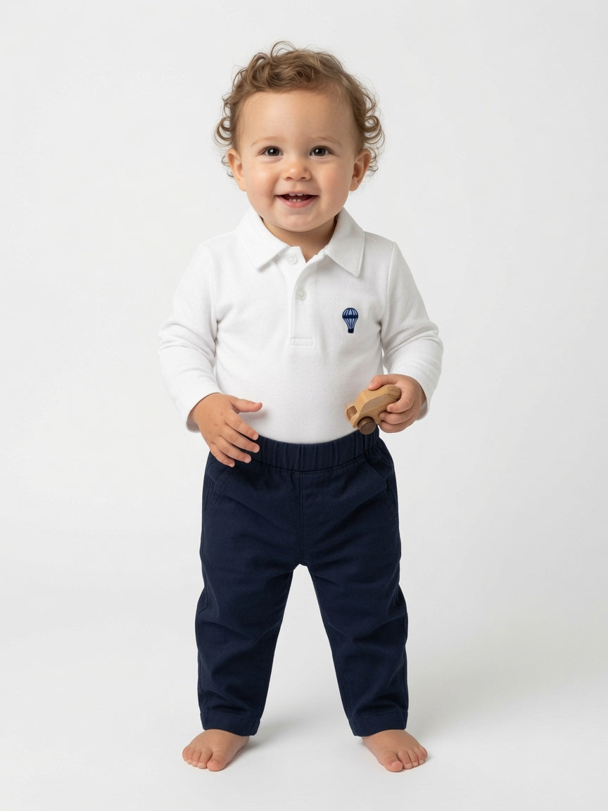 A toddler standing barefoot against a light gray studio background, wearing a white long-sleeve polo shirt with a small blue hot air balloon logo on the left chest and dark navy pants.