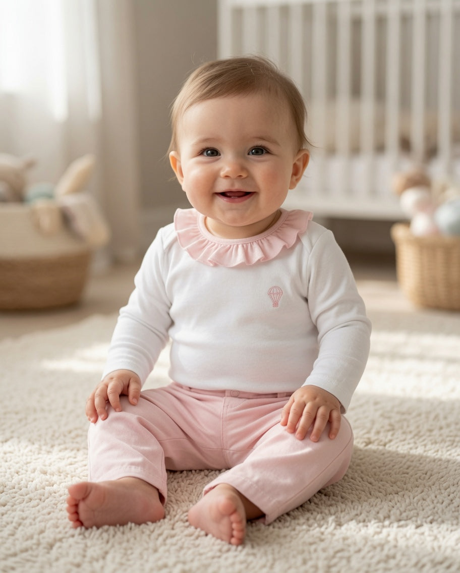 A smiling baby sitting on a cream carpet in a softly lit nursery, wearing a white long-sleeve top with a pink ruffled collar and pink pants.