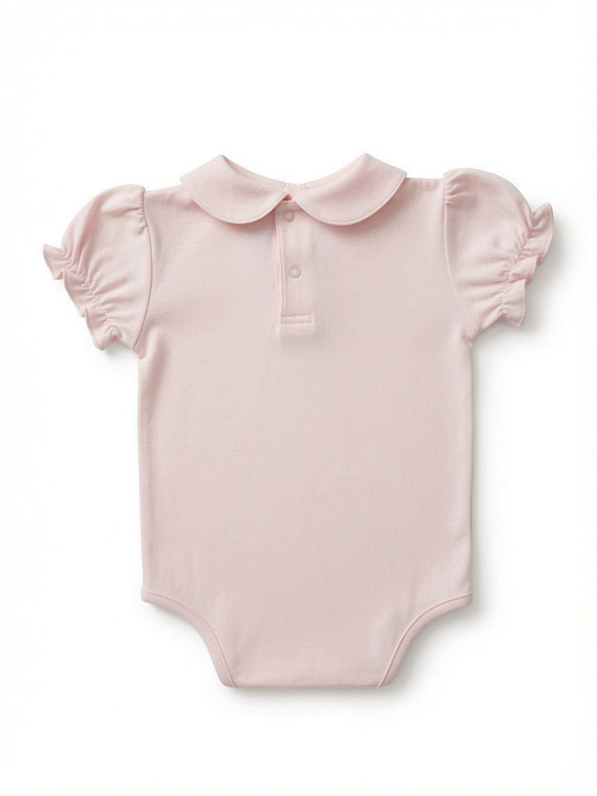 A light pink baby bodysuit with a Peter Pan collar, short ruffled sleeves, and a small embroidered hot air balloon logo on the left chest made from Peruvian Pima Cotton on the back side.