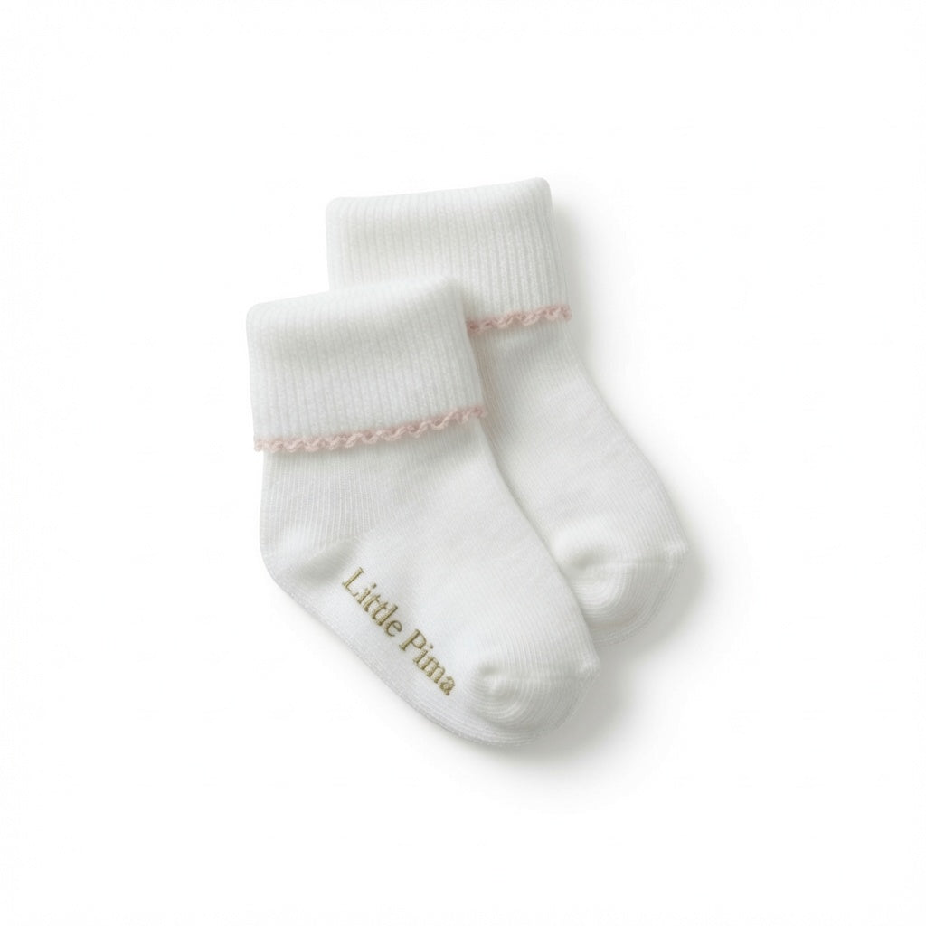 A pair of white baby socks with a delicate pink ruffled trim at the ribbed cuffs and the brand name 'Little Pima' printed in soft sage green on the sole, shown on a clean white background.