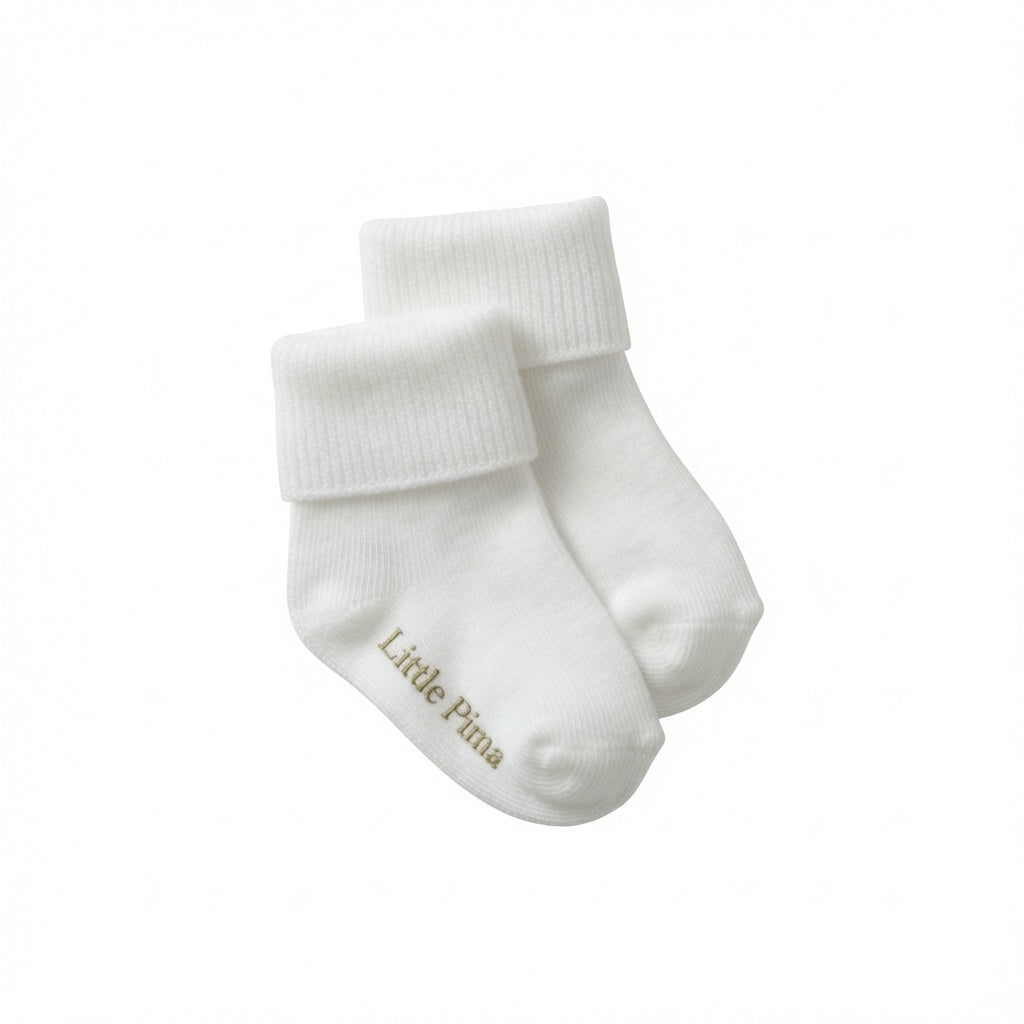 A pair of white baby ankle socks with the brand name 'Little Pima' printed in soft sage green on the sole, shown against a plain white background.
